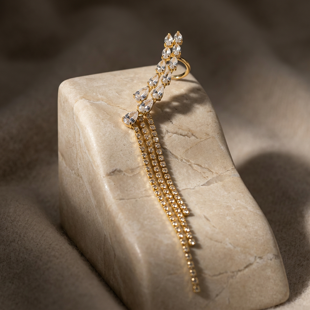 A gold-toned statement earring with clear crystal-like stones, draped over a beige stone block on a soft taupe fabric background.