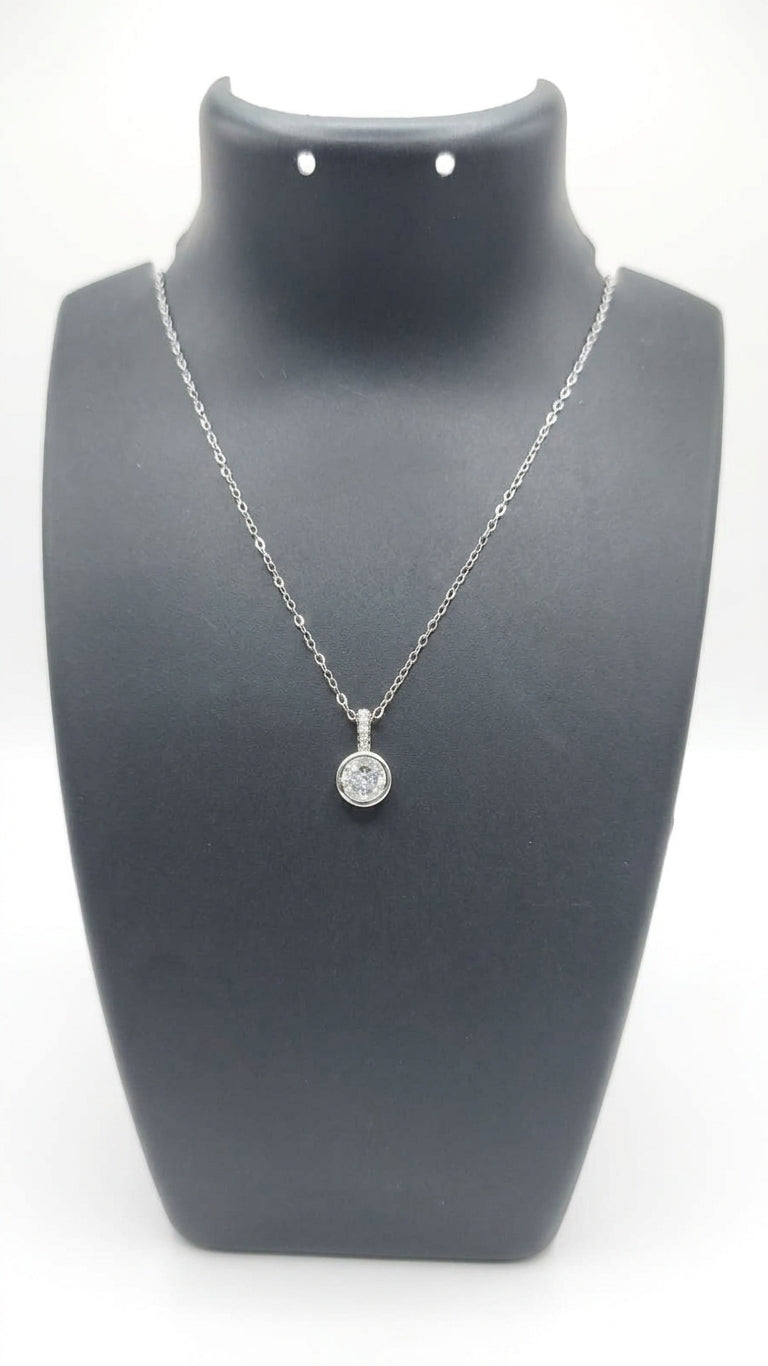 Woman wearing silver bezel-set round CZ solitaire pendant necklace on fine cable chain at collarbone in black v-neck top against warm beige background – diamond pendant necklace AureliaBlends