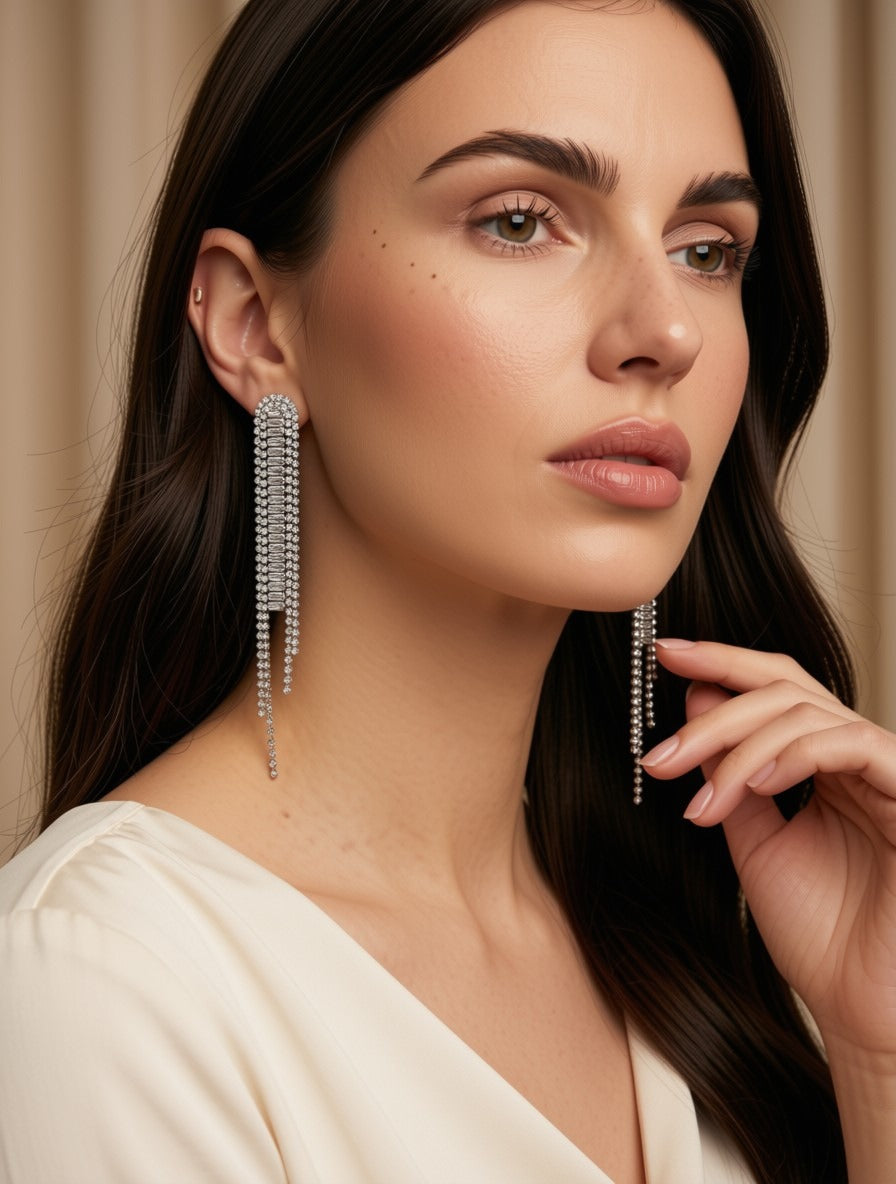 A close-up of a woman wearing long, dangling silver beaded earrings with multiple strands hanging from oval studs, against a soft beige background.
