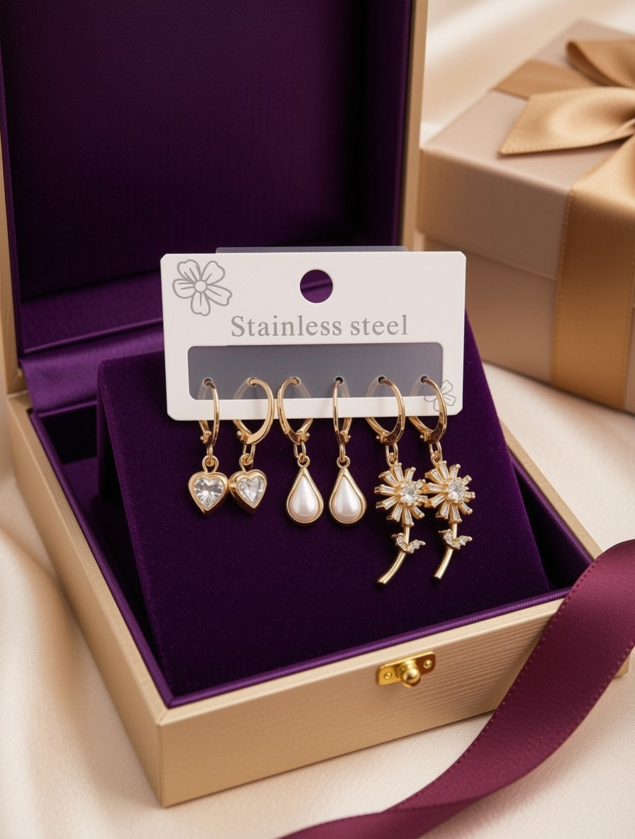 A set of five gold-tone stainless steel dangle earrings displayed on a white card inside an open purple jewelry box. The earrings include a heart, pearl teardrop, and floral designs with clear stones.