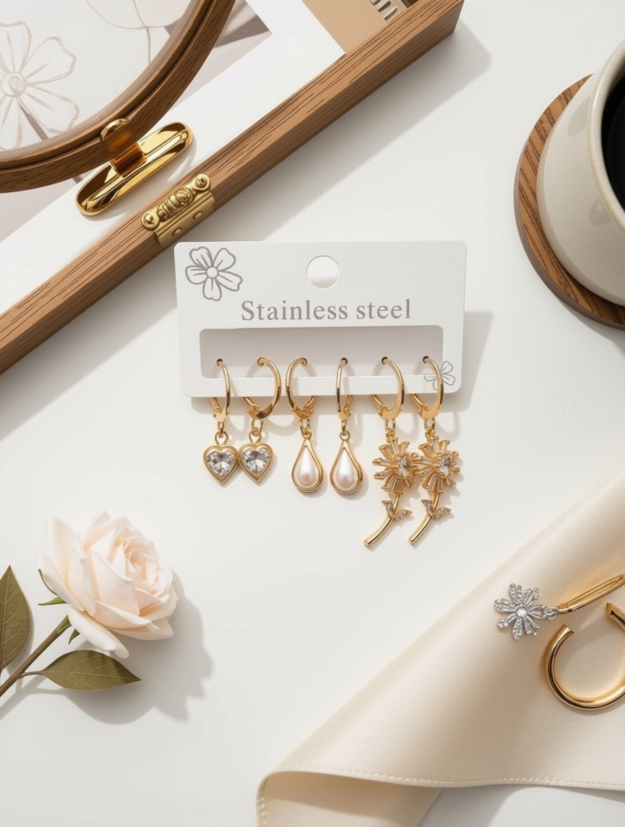 A set of five gold-tone stainless steel dangle earrings displayed on a white card labeled 'Stainless steel,' arranged on a light tabletop with decorative props including a rose, folded cream fabric, a wooden tray, and a cup.