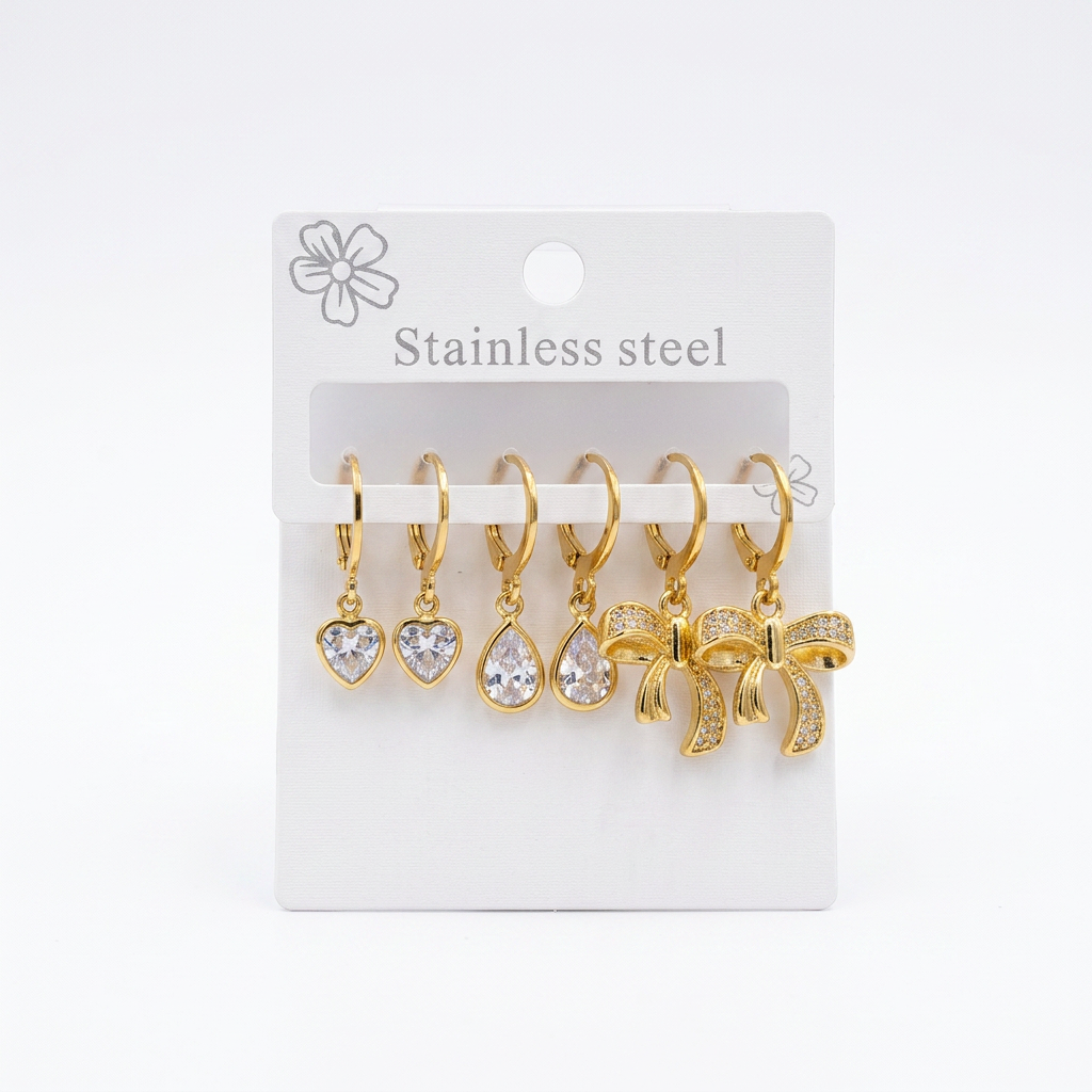 A set of five gold-tone stainless steel earrings on a white display card. The earrings include heart, teardrop, and bow-shaped clear crystal pendants.