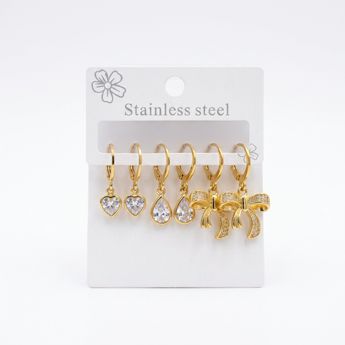 A set of five gold-tone stainless steel earrings on a white display card. The earrings include heart, teardrop, and bow-shaped clear crystal pendants.