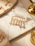 Four gold-tone dangle earrings displayed on a white card labeled 'Stainless steel,' arranged on a cream marble surface with warm beige props and jewelry accessories around them.