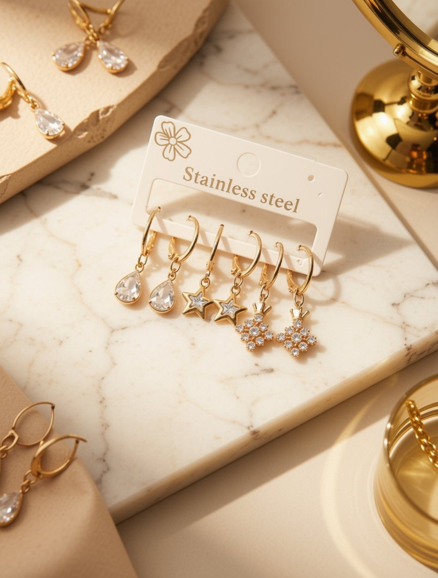 Four gold-tone dangle earrings displayed on a white card labeled 'Stainless steel,' arranged on a cream marble surface with warm beige props and jewelry accessories around them.