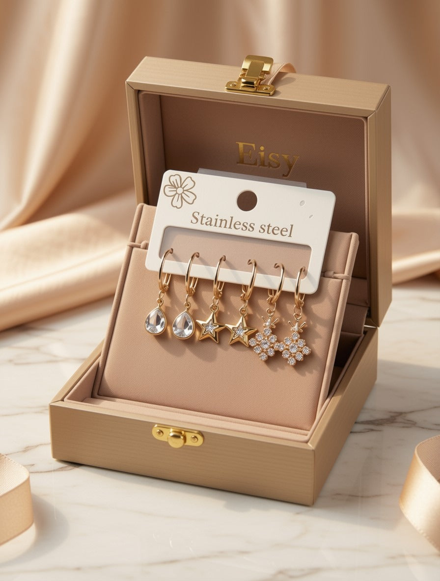 A set of five gold-tone dangle earrings displayed in an open beige jewelry box. The earrings include clear teardrop, star, and snowflake designs on stainless steel hooks, attached to a white card labeled 'Stainless steel' with 'Etsy' printed inside the box.