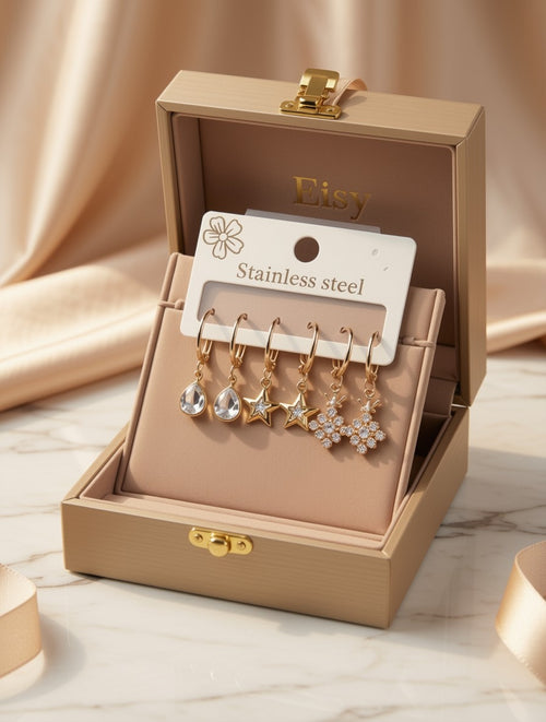 A set of five gold-tone dangle earrings displayed in an open beige jewelry box. The earrings include clear teardrop, star, and snowflake designs on stainless steel hooks, attached to a white card labeled 'Stainless steel' with 'Etsy' printed inside the box.