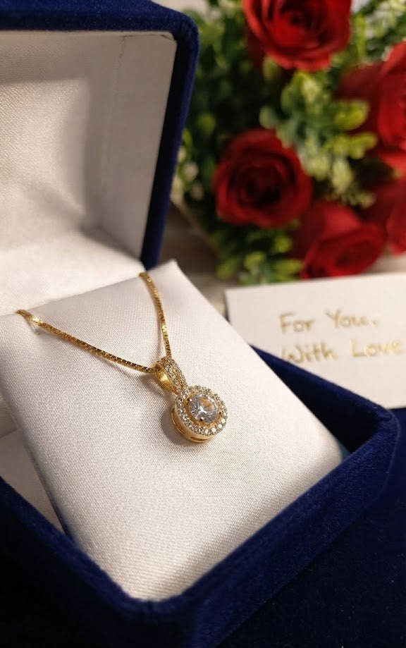 Gold round halo CZ diamond solitaire pendant necklace on black jewellery bust surrounded by vibrant jewel-toned flowers, peacock feathers, amethyst crystals and For You With Love card – UAE luxury halo necklace AureliaBlends