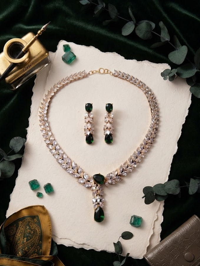 Emerald green CZ crystal leaf necklace and drop earrings set flat lay on white paper with green gems and eucalyptus – AureliaBlends