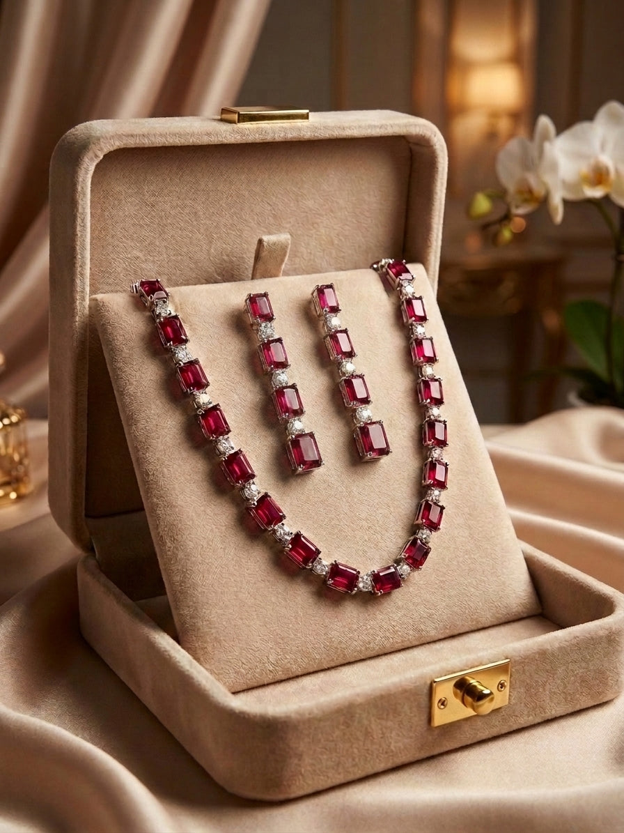 Indian woman wearing ruby red emerald cut CZ tennis necklace and long drop earrings in black dress with cream satin background – AureliaBlends