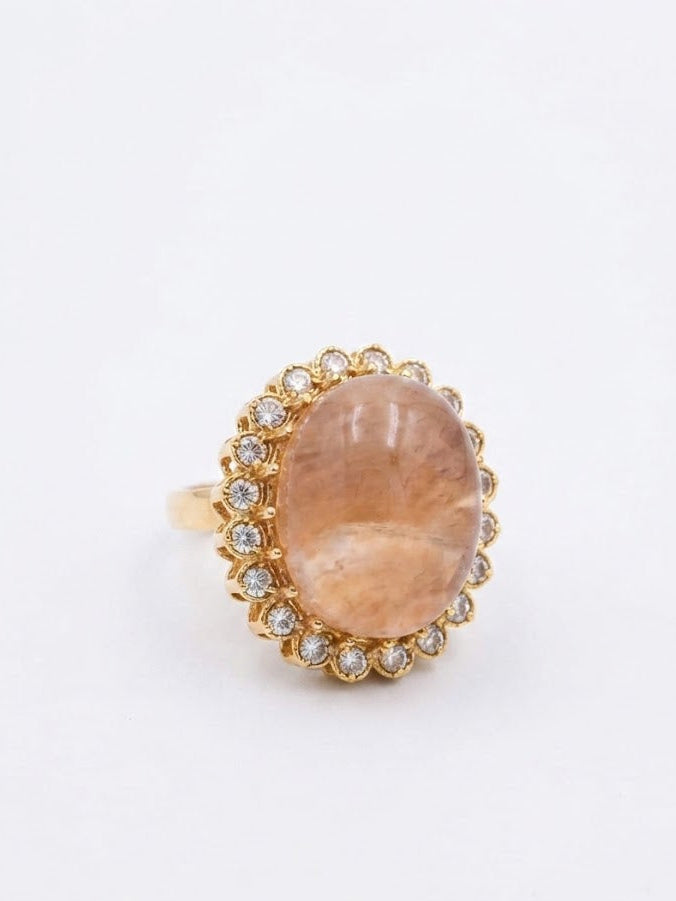 Gold oval peach stone halo cocktail ring flat lay on cream linen fabric with gold jewelry box sunglasses pearl earrings and lavender sprigs – AureliaBlends