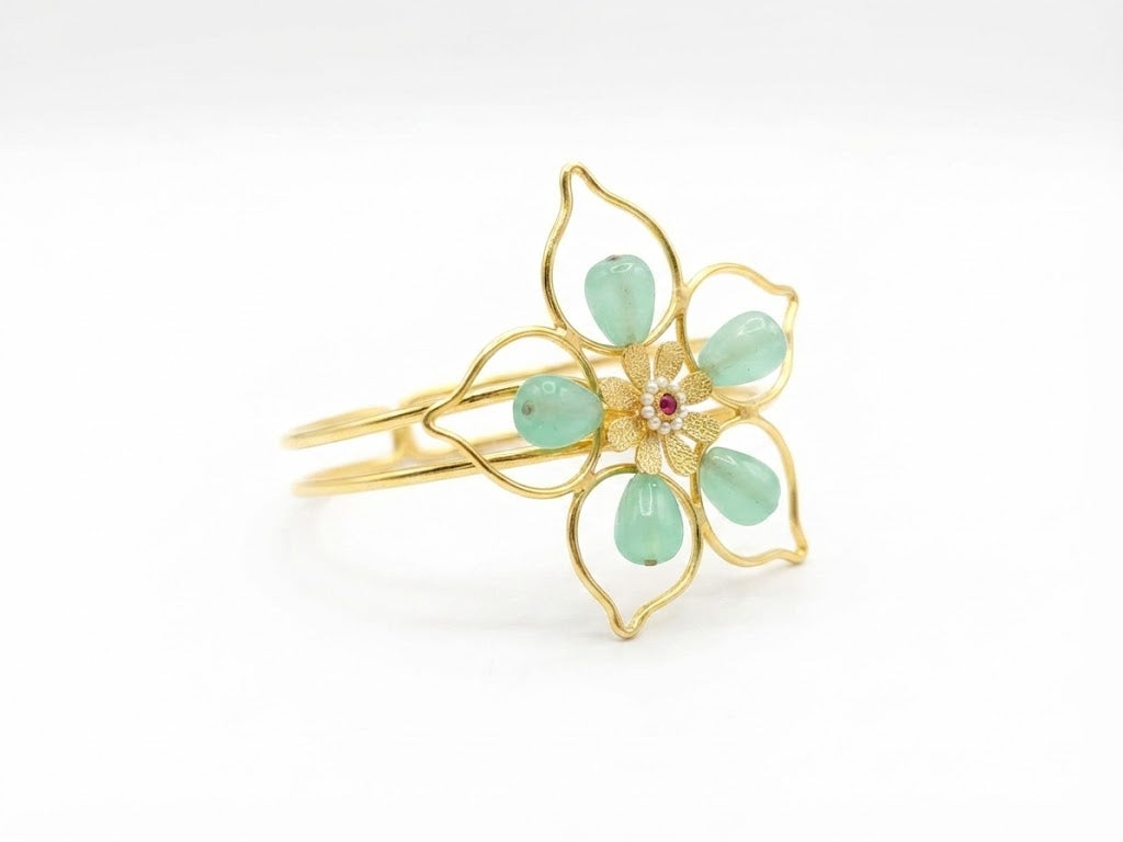 Gold open cuff bangle bracelet with green aventurine teardrop stone floral motif and ruby CZ centre displayed on beige velvet jewelry cushion in warm jewelry store lighting – AureliaBlends