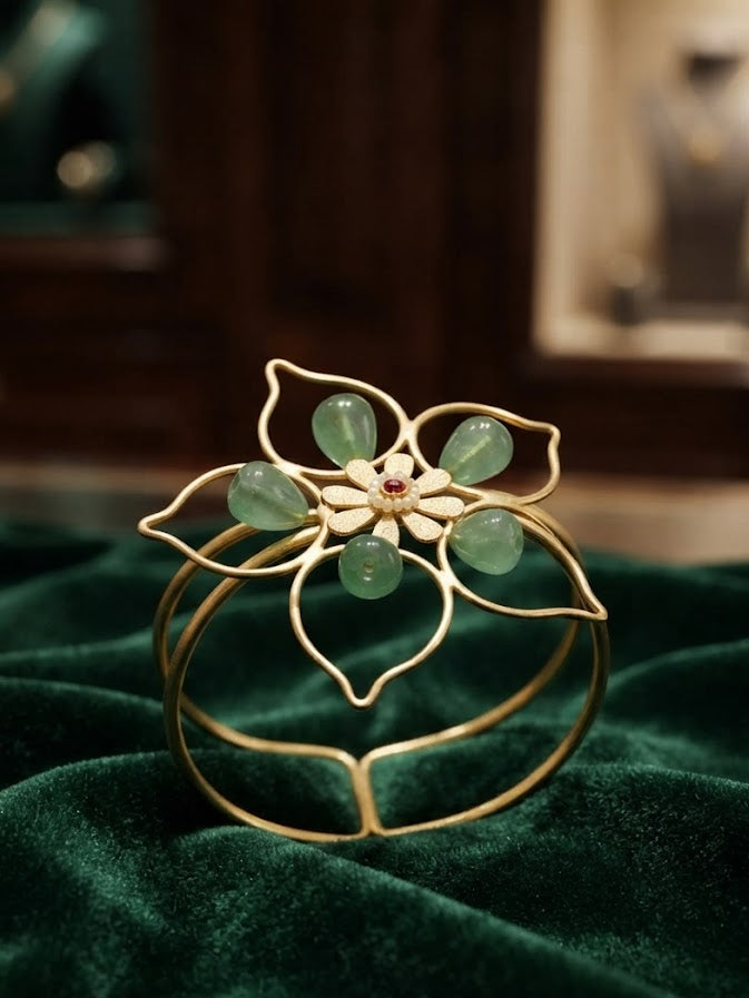 Gold open cuff bangle bracelet with green aventurine floral motif and ruby CZ centre displayed on dark green velvet fabric in jewelry store setting – AureliaBlends