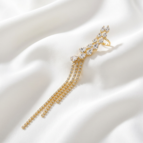 A gold-toned statement ring featuring a cluster of clear teardrop and marquise-cut stones with three dangling chain strands, displayed on draped white satin fabric.
