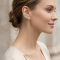 A close-up side profile of a woman wearing a delicate gold ear cuff with clear rhinestones and long dangling chain earrings, against a soft neutral background.
