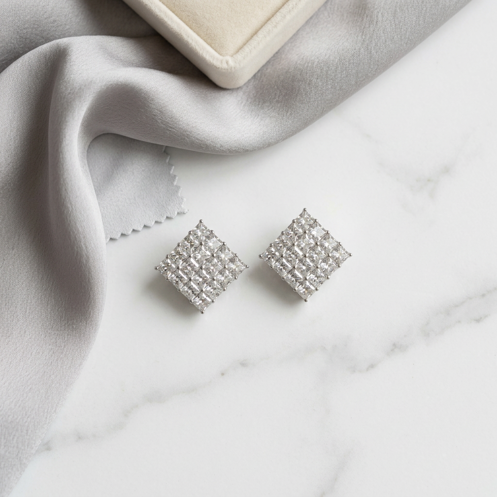 A pair of square stud earrings covered in small clear stones, arranged on a white marble surface with a draped light gray fabric and part of a cream jewelry box visible.