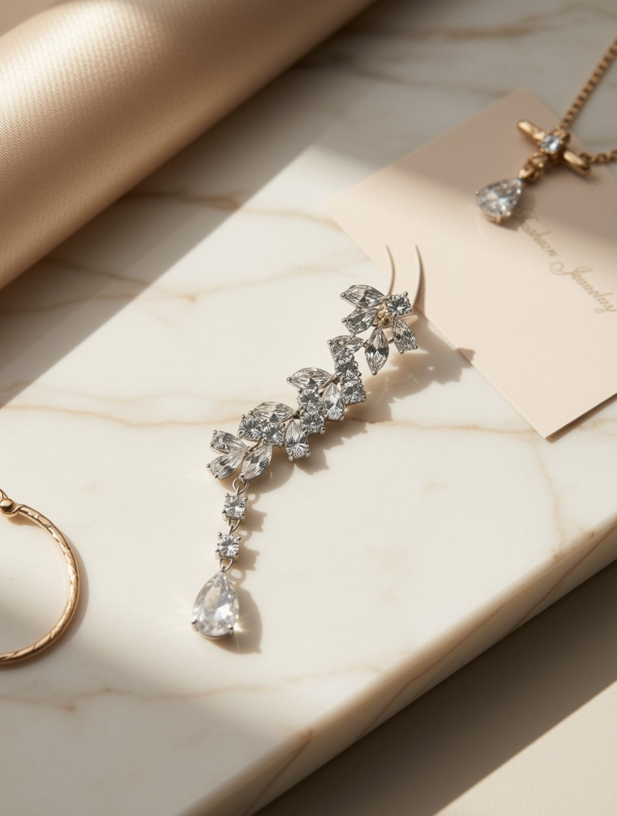A silver-toned floral drop earring with clear teardrop crystals, displayed on a cream marble block with soft warm lighting and a jewelry card nearby.