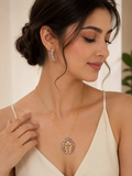 A woman wearing a gold pendant necklace and matching drop earrings with pink and purple stones, styled with a cream-colored spaghetti strap top.