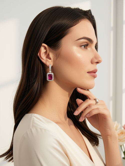A woman in profile wearing a dangling earring with a rectangular pink gemstone and a silver-toned setting, paired with a silver ring on her finger. She is wearing a cream-colored top against a bright, softly lit background.
