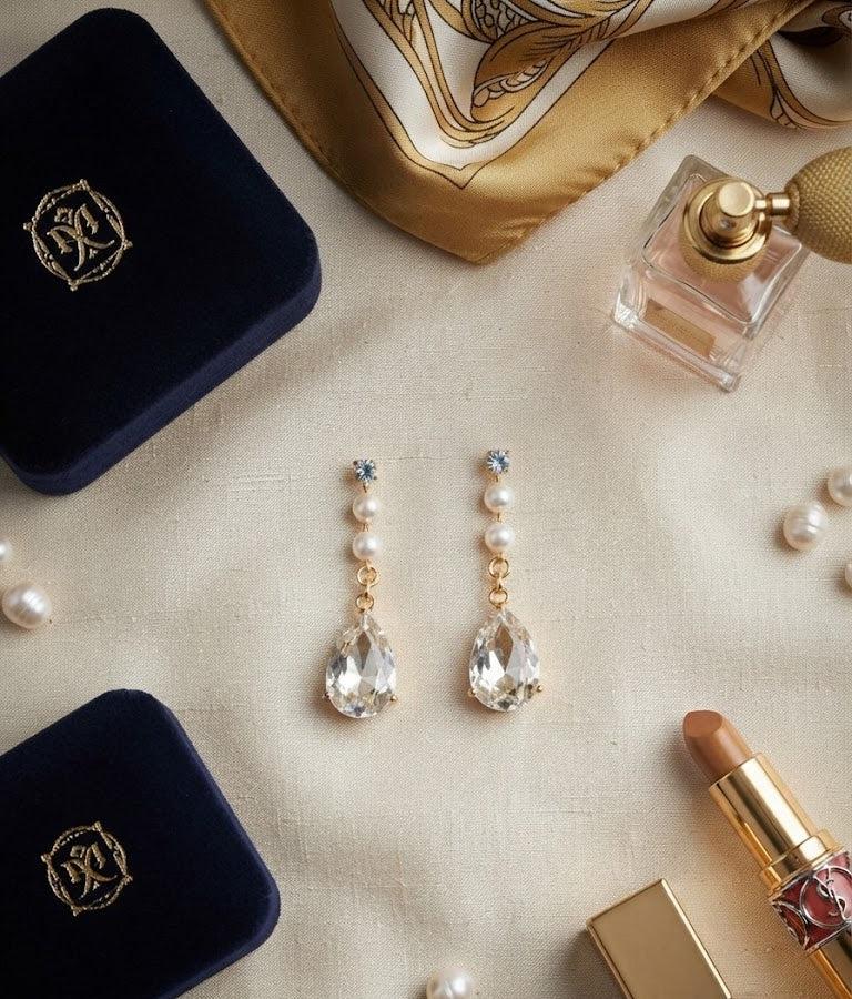Gold CZ pearl chain teardrop crystal drop earrings editorial flat lay on cream silk with navy velvet gift boxes, perfume, lipstick and scattered pearls – AureliaBlends