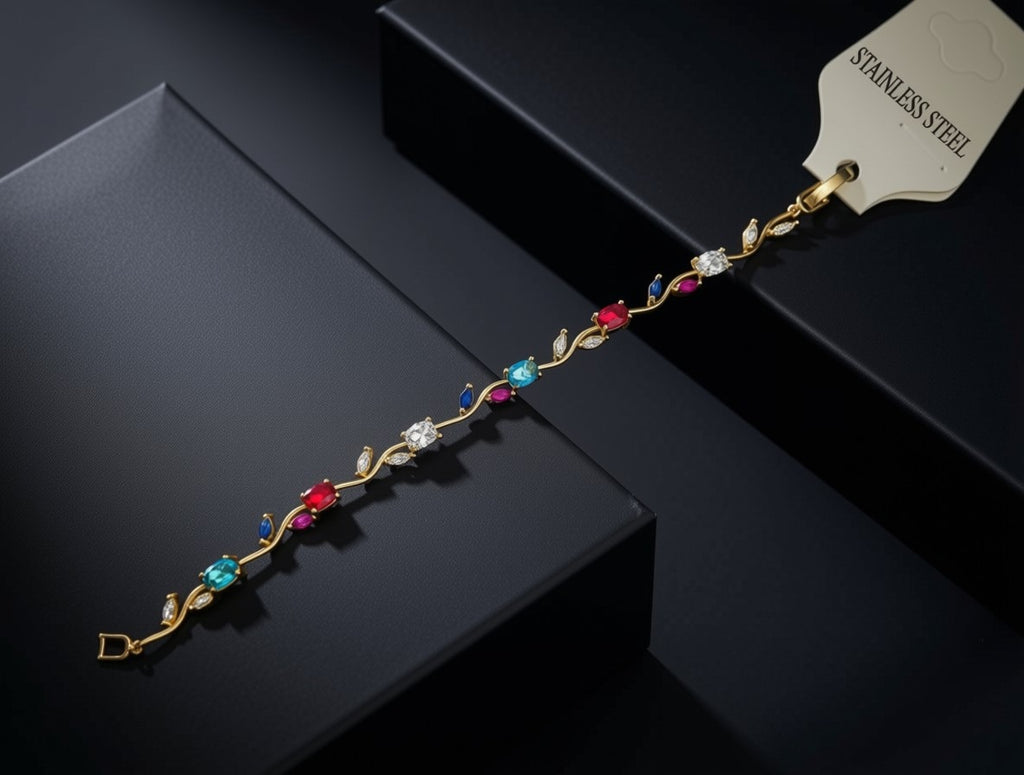 A gold-toned stainless steel bracelet with colorful gemstone accents, displayed diagonally on a dark gray box with a white tag reading 'STAINLESS STEEL'.