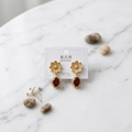 A pair of gold flower-shaped drop earrings with oval dark red gemstones, displayed on a white card labeled 'Italian Accessories' on a white marble surface with small decorative stones and dried flowers.