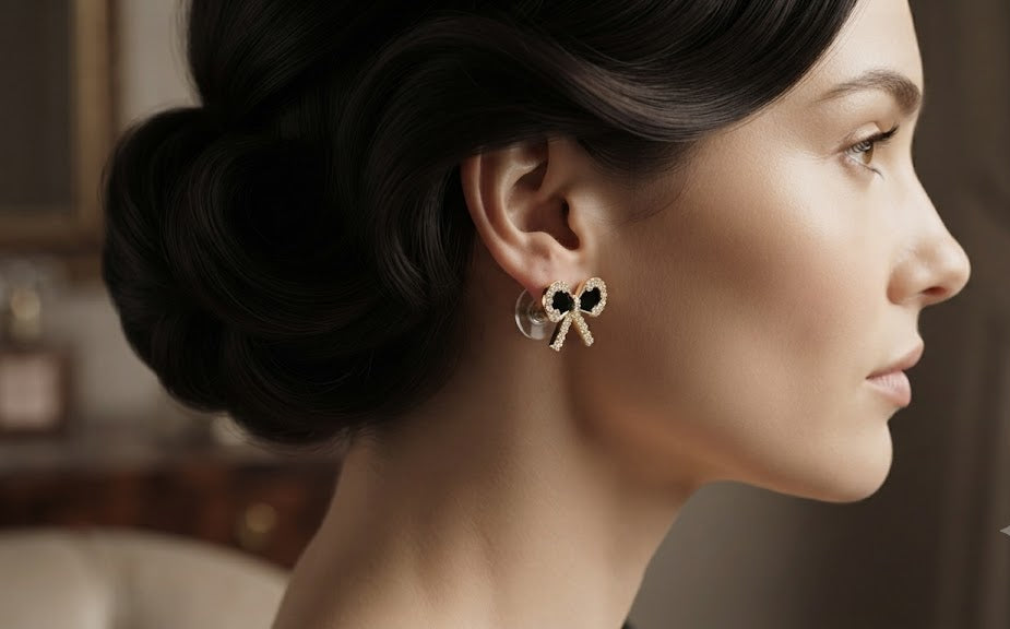 Gold pavé CZ black bow stud earrings on a white velvet ring cushion surrounded by a gold bangle, vintage compact, eucalyptus leaves and a Timeless Elegance card on blush linen – AureliaBlends
