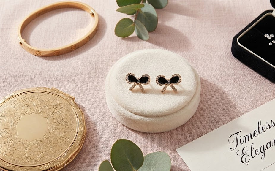 Gold pavé CZ black bow stud earrings on a white velvet cushion with a gold bangle, vintage compact, eucalyptus, a gift box with gold ribbon and Timeless Elegance card on black velvet – AureliaBlends