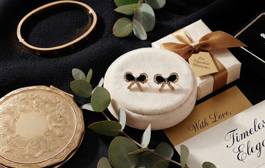 Gold pavé CZ black bow stud earrings on a white velvet cushion surrounded by blush and burgundy roses, lavender, eucalyptus, a gold bangle, vintage compact and a For Timeless Moments gift box – AureliaBlends