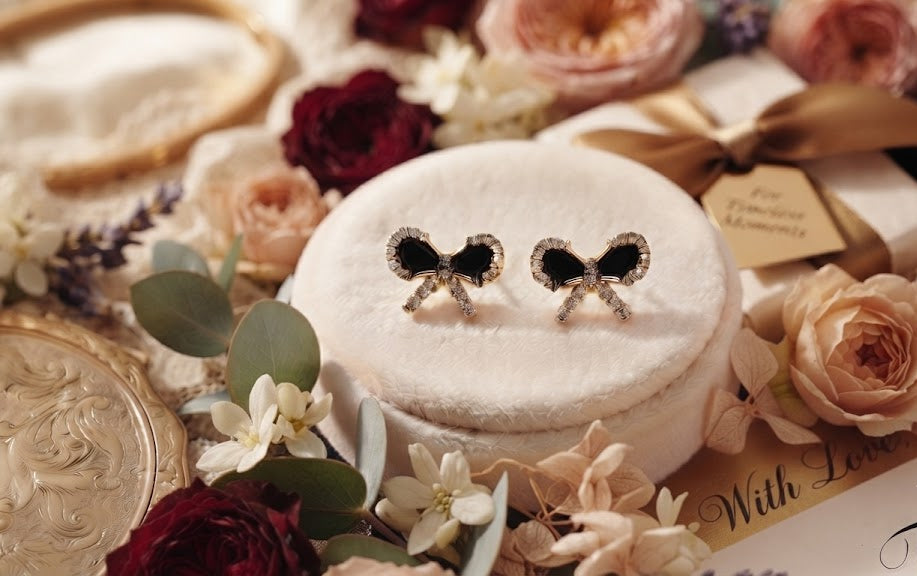 Gold pavé CZ black bow stud earrings on a white velvet cushion surrounded by blush and cream roses, white florals, eucalyptus and a For Timeless Moments gift box – AureliaBlends Timeless Elegance Collection