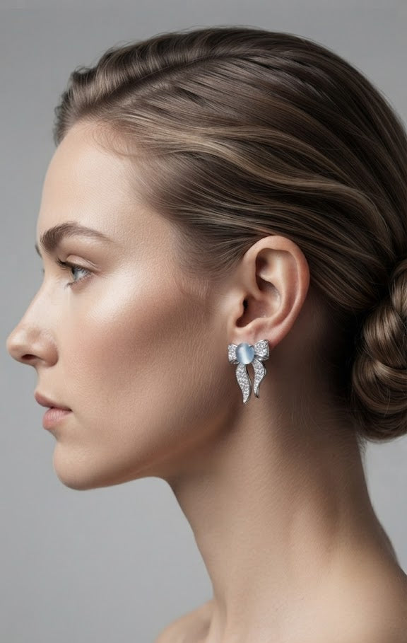 Silver pavé CZ moonstone bow stud earrings flat lay on white marble with a blue velvet pouch, blue paisley silk scarf, vintage leather journal, white orchid, silver fountain pen and blue aviator sunglasses – AureliaBlends