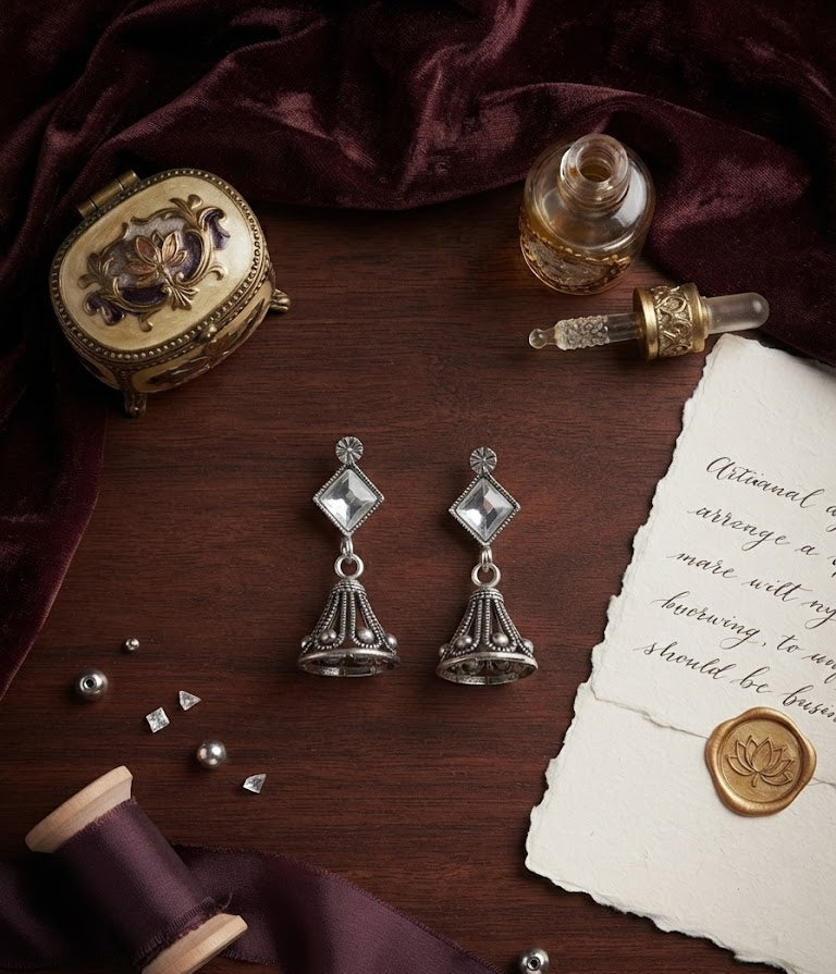 Oxidised silver diamond CZ and open filigree jhumka bell drop earrings flat lay on dark walnut wood with vintage gold trinket box, perfume bottle, lotus wax seal, plum ribbon and scattered CZ crystals – AureliaBlends luxury earrings