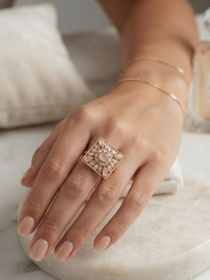 Woman wearing rose gold square filigree cocktail statement ring with pink opal and CZ stones on index finger with neutral manicure on white marble surface with perfume bottle in background – AureliaBlends