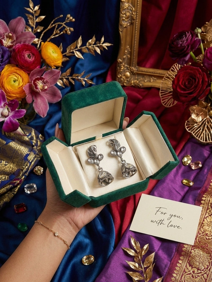 Model with loose dark hair wearing oxidised silver peacock CZ kundan and jhumka bell drop earrings surrounded by blush pink peach and cream roses ranunculus and fern leaves with For You With Love card – AureliaBlends romantic bridal earrings