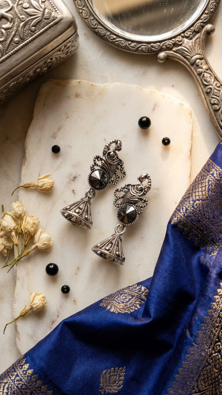 Oxidised silver peacock scroll and hematite rivoli jhumka bell drop earrings on hammered gold thali tray with scattered hematite crystals and pearl beads on dark burgundy velvet – AureliaBlends statement earrings
