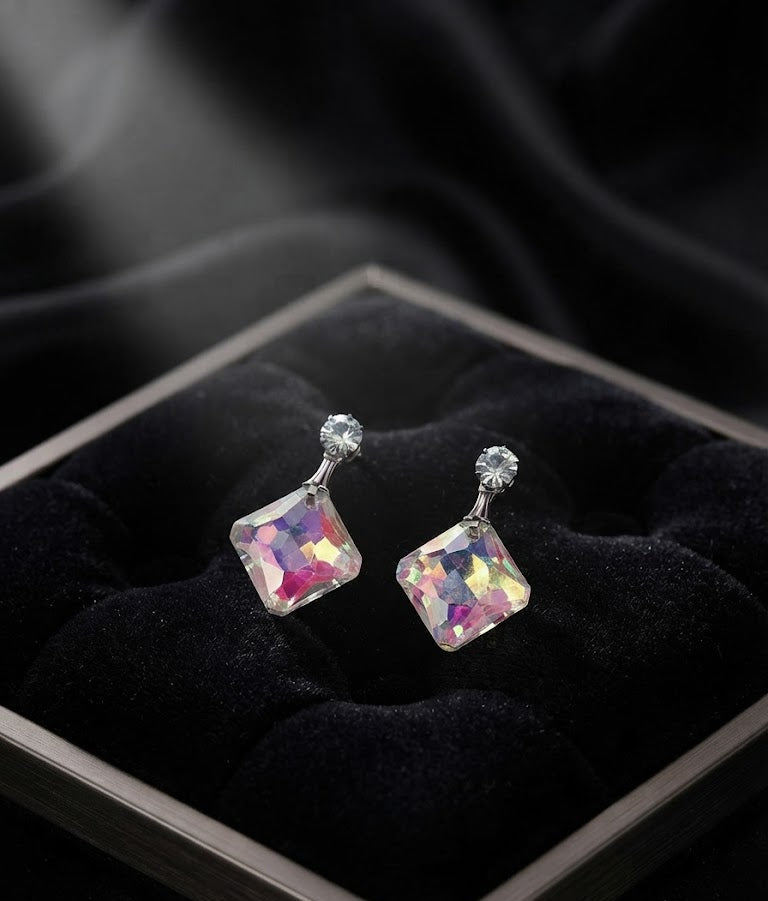Silver CZ stud and aurora borealis iridescent square crystal drop earrings displayed on sculptural gunmetal silver earring stand on black marble geometric base with gold inlay lines against jewel-toned velvet background – AureliaBlends editorial