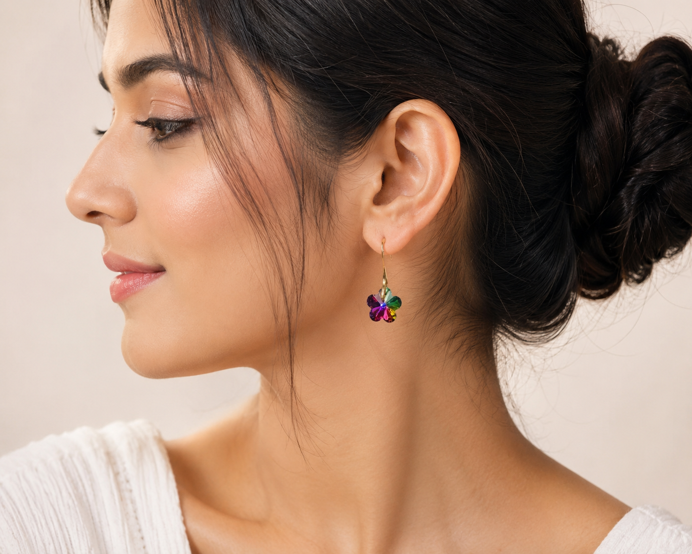 Model with elegant low bun wearing gold-plated vitrail rainbow 5-petal flower crystal hook drop earrings showing multicolour magenta emerald violet ruby blue shimmer against warm beige background – AureliaBlends