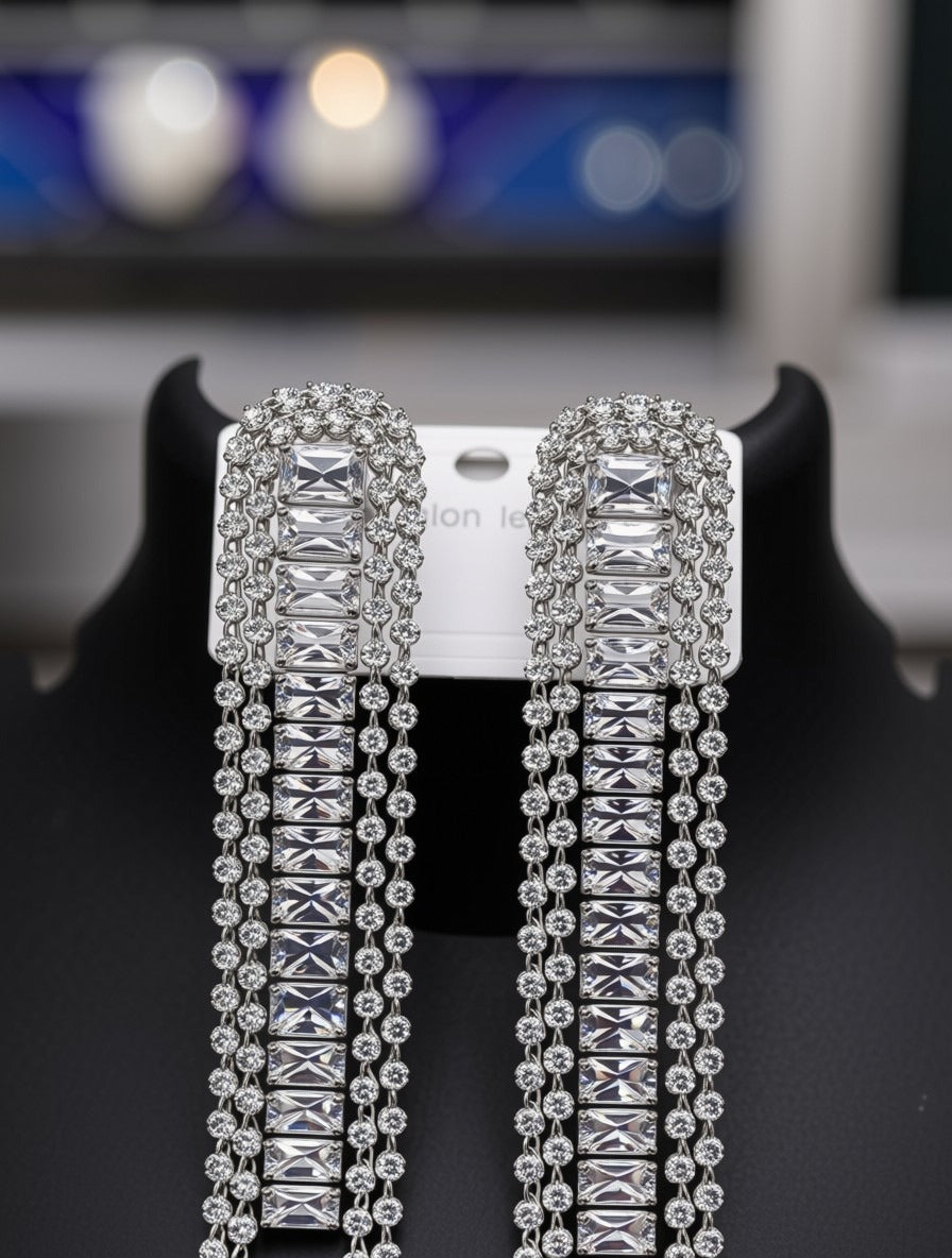 A pair of long silver-toned statement earrings with rectangular clear rhinestones and dangling chain fringe, displayed on a black jewelry stand with a white card backing.