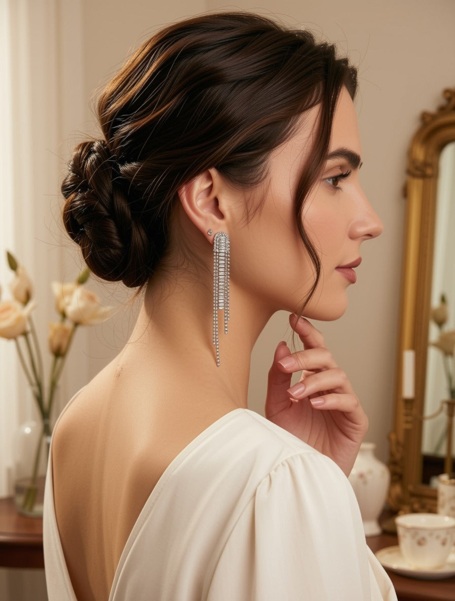 A woman in profile wearing a long, sparkling silver dangle earring with clear stones, styled in an elegant updo, wearing a cream off-the-shoulder dress in a softly lit room.