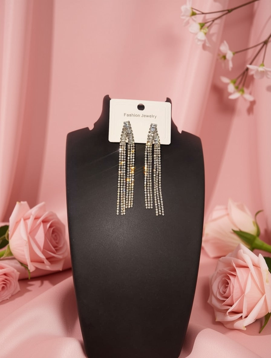 A pair of long dangling statement earrings displayed on a black jewelry bust against a pink draped backdrop with pink roses around it. The earrings have silver-toned metal with rows of clear rhinestones and small gold-toned bead accents.