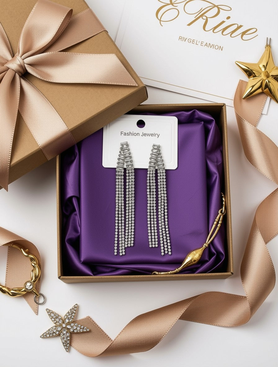 A pair of silver rhinestone tassel drop earrings displayed on a white card inside an open brown gift box lined with purple satin. The box is styled with a matching brown ribbon, a gold star ornament, and a gold bracelet on a white background.