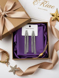 A pair of silver rhinestone tassel drop earrings displayed on a white card inside an open brown gift box lined with purple satin. The box is styled with a matching brown ribbon, a gold star ornament, and a gold bracelet on a white background.