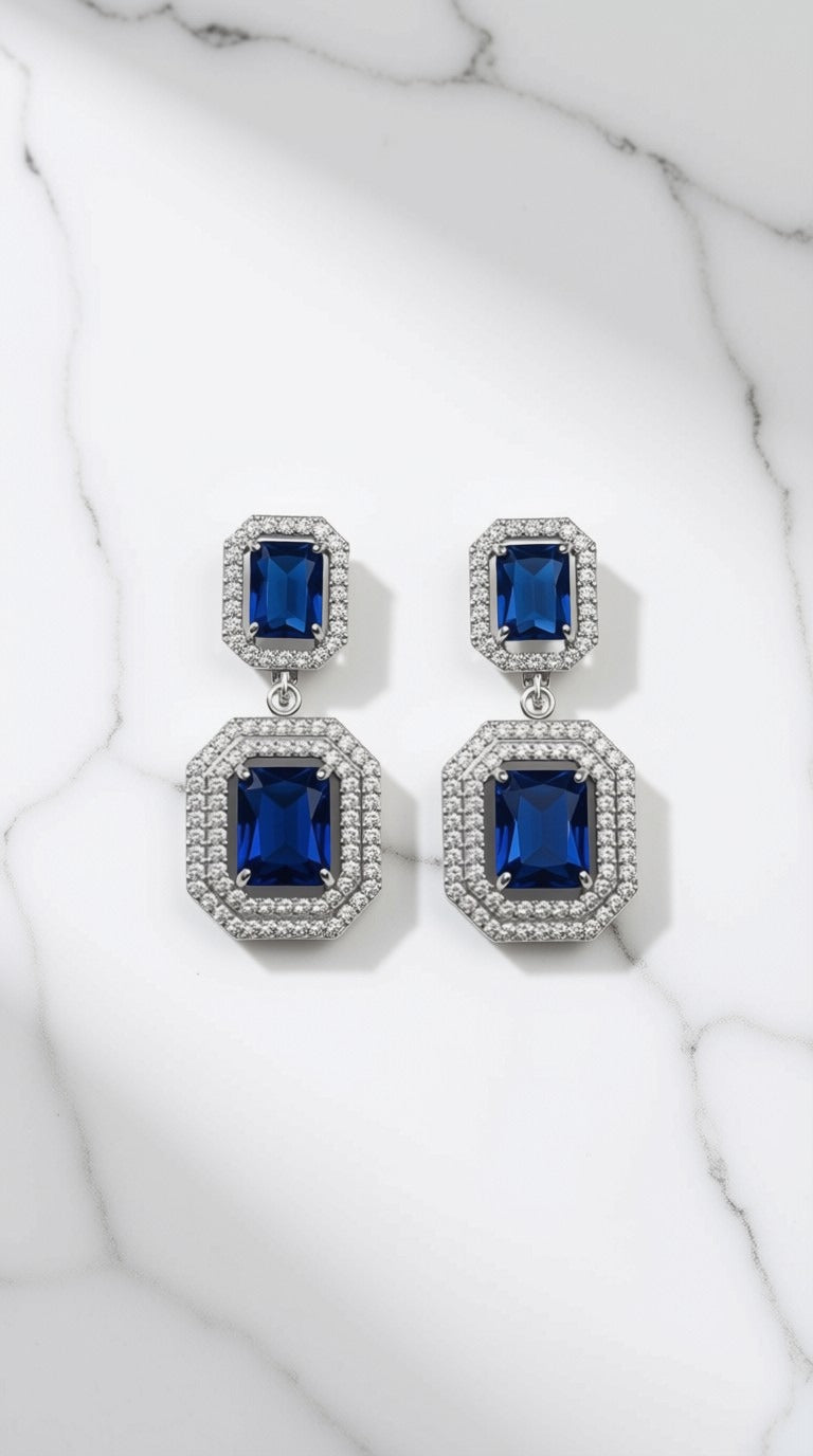 A pair of silver-toned drop earrings with rectangular blue gemstones surrounded by small clear stones, displayed on a white marble surface.