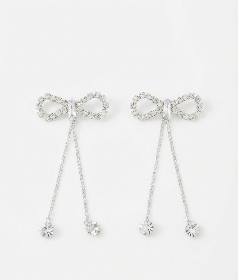 Silver CZ open bow tassel chain drop earrings on a navy blue velvet jewellery pouch on a dark black background with scattered loose CZ stones and a silver satin ribbon tag – AureliaBlends dark editorial