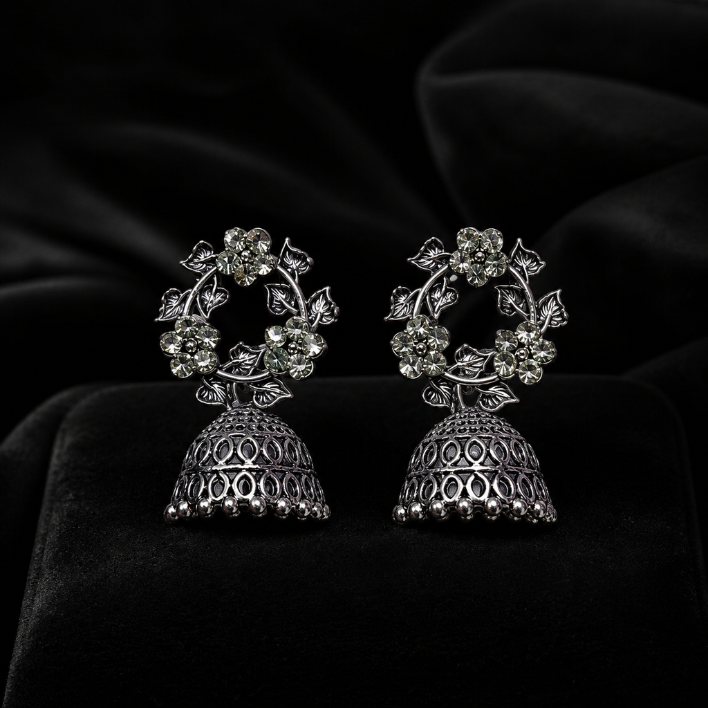 AureliaBlends oxidised silver CZ floral wreath and open oval lattice jhumka bell drop earrings lifestyle editorial flat lay on cream satin with white dahlia gypsophila and gold-rimmed dish