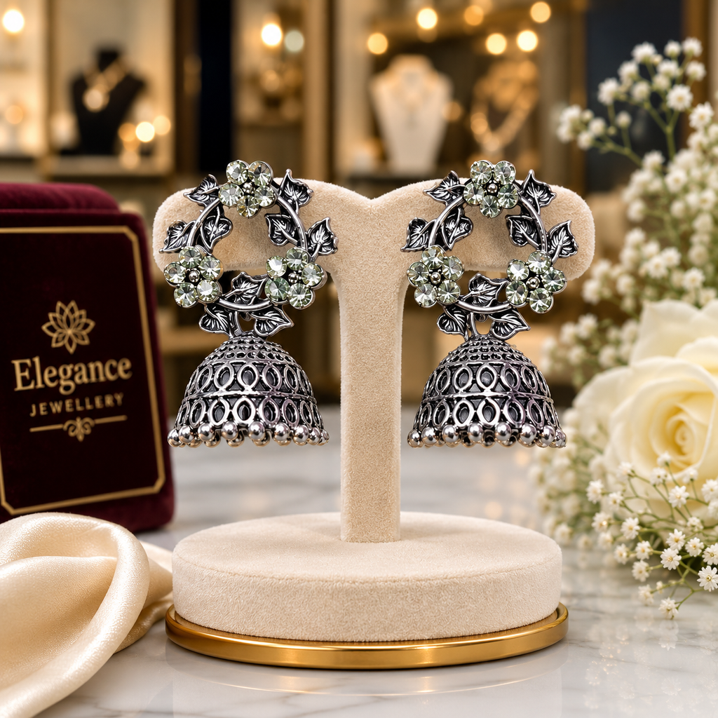 Oxidised silver CZ floral wreath and open oval lattice jhumka bell drop earrings editorial boutique shot on cream suede T-bar stand with warm bokeh lighting white roses gypsophila and Elegance Jewellery branding – AureliaBlends antique silver earrings