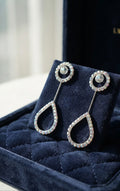 Silver CZ round halo solitaire chain teardrop pavé long drop earrings flat lay on white marble with scattered iridescent crystal bicone beads, a navy blue jewellery box and a silver fountain pen with a champagne silk scarf – AureliaBlends lifestyle shot