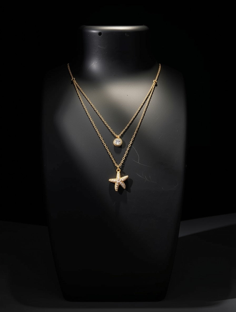 A double-strand gold necklace displayed on a black mannequin bust against a black background. The necklace features a delicate chain with a small round white stone pendant and a longer chain with a starfish-shaped pendant.